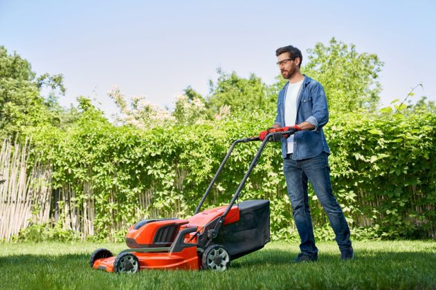 Beau jardinier masculin en gants qui coupe le gazon avec une tondeuse électrique dans le jardin au jour.